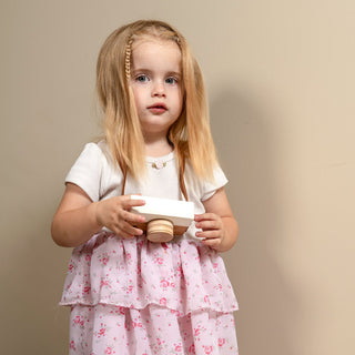A young girl with blonde hair stands on a beige carpet against a beige wall, wearing a white short-sleeve top and a pink tiered skirt with a small floral print, paired with pink socks.