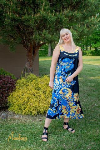 Image of a beautiful young woman with long blond hair wearing a decorative summer dress with a patriotic Ukrainian design. Image of a blue-yellow decorative bird and bright yellow-blue sunflowers on a black background and the slogan "Standing with Ukraine"