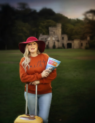 Woman in red hat and sweater holding a book and suitcase in a grassy field with ruins in the background