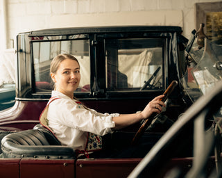 A smiling woman with curly hair sits in a vintage car, holding a wooden steering wheel. The background is a softly blurred indoor setting with warm lighting.