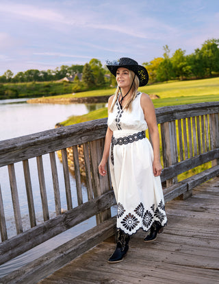 Woman in a white dress with black patterns and a black hat standing on a wooden bridge by a lake.