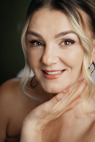 A close-up portrait of a smiling woman with dark brown hair highlighted with blonde streaks, wearing dangling earrings against a soft olive green background.
