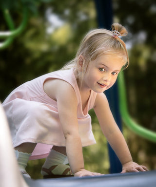 A young blonde girl in a light pink sleeveless dress and diaper cover crawls on a playground slide outdoors, with a blurred green park background.