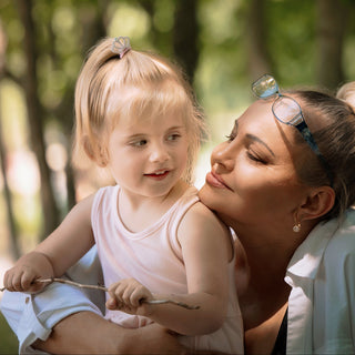 A woman in a white robe looking upward while a young blonde girl in a pale pink dress gently touches her hair, set against a softly blurred outdoor forest background.
