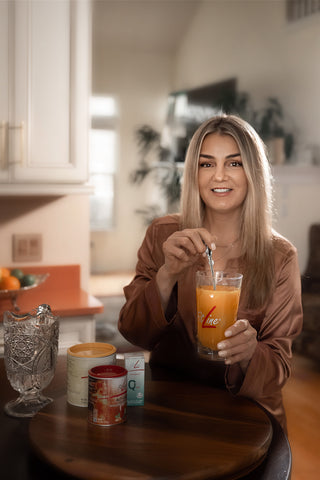A woman with blonde hair wearing a brown satin shirt sits at a wooden table in a kitchen, holding a glass of orange juice with a metal straw. The glass features the red and white 'Zine' logo.
