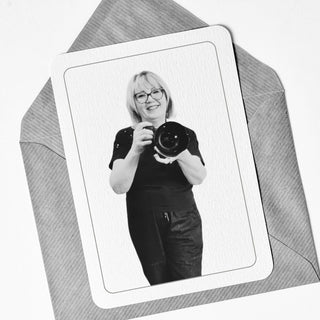 Black and white photo of a person holding a camera, framed as if on a card, with a textured gray background.