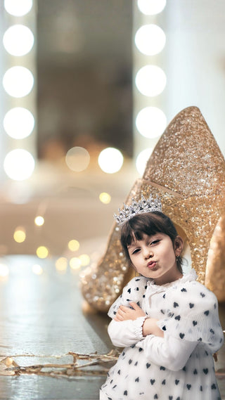 Young girl in a white dress with black heart patterns and a tiara, standing in front of a blurred festive background with lights.