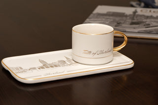 Ceramic cup with gold handle on a matching tray on a dark surface with a newspaper in the background.