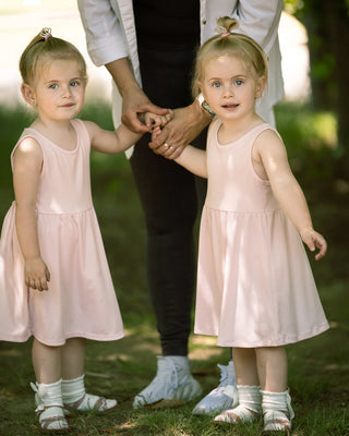Two young girls wearing matching pale pink sleeveless dresses stand outdoors holding an adult's hand. The scene is set on grass with blurred green foliage in the background.