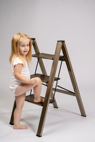 A young girl with long blonde hair wearing a white bodysuit sits on a three-step wooden step ladder with a metallic bronze finish, photographed against a seamless light gray studio background.