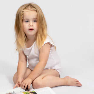 A young blonde girl in a white sleeveless dress sits cross-legged on a white studio floor, looking at an open magazine.