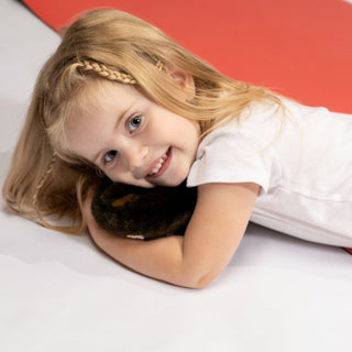 A young girl with blonde hair lies on a red exercise mat on a white studio floor, wearing a white short-sleeve bodysuit. A small dumbbell is visible near the top right of the mat.