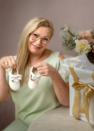 A woman with blonde hair and glasses holds a pair of tiny white baby booties next to a large white gift box with gold ribbon and tissue paper, with a floral arrangement in the background.