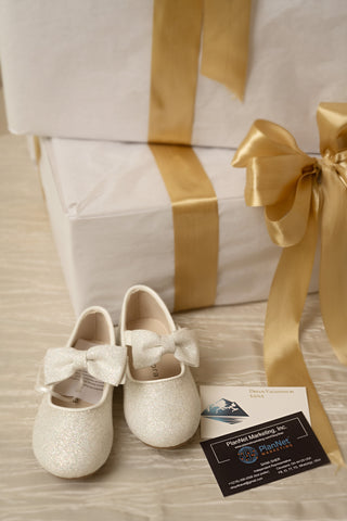 A pair of white glitter baby shoes with bow details, displayed on a gift box next to a large gold satin bow and ribbon, with two business cards in the foreground on a light wood surface.