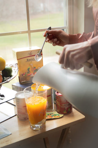 A person pouring hot water from a white pitcher into a glass of orange juice on a wooden table, with tea boxes and a window in the background.