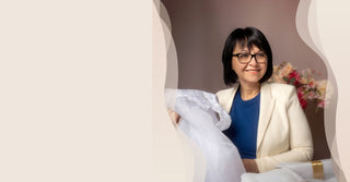 A woman with short black hair and glasses, wearing a cream blazer over a blue top, holds up a white lace bridal veil or dress while seated in an indoor setting with a gift box in the foreground.