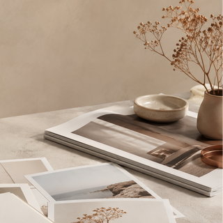 A styled interior tabletop scene with a ceramic mug of coffee, scattered photo prints, an open book or magazine, small bowls, and dried flowers in a vase against a warm beige wall.