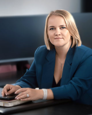 A woman with blonde hair sits at a desk in an office environment, wearing a teal blazer over a black top. She is holding a pen and writing in an open notebook.