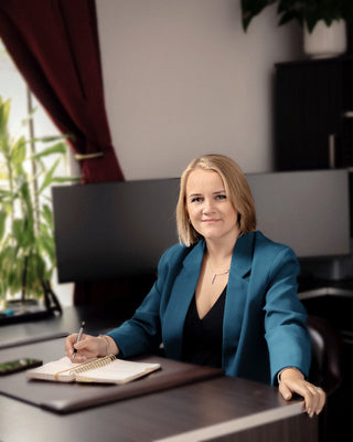Woman in a blue blazer sitting at a desk with a notebook and pen, in an office setting.